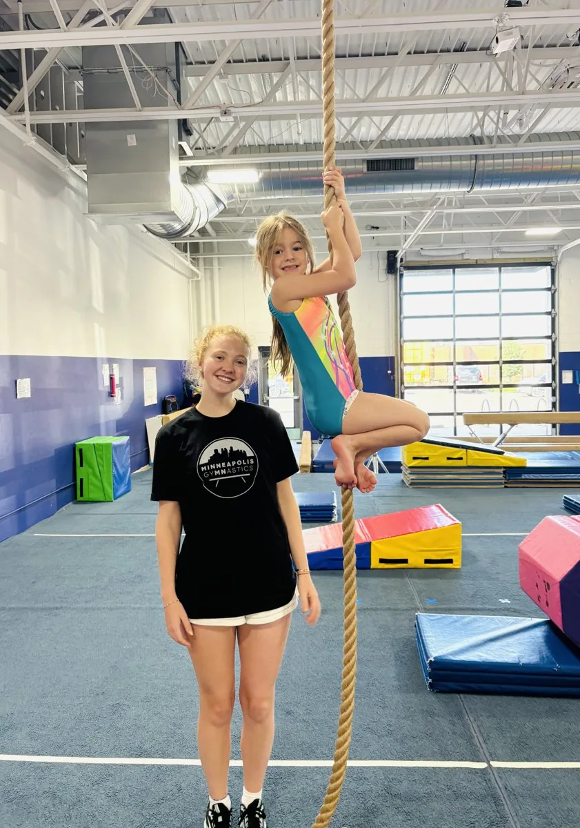 A smiling coach stands beside a young gymnast climbing the rope