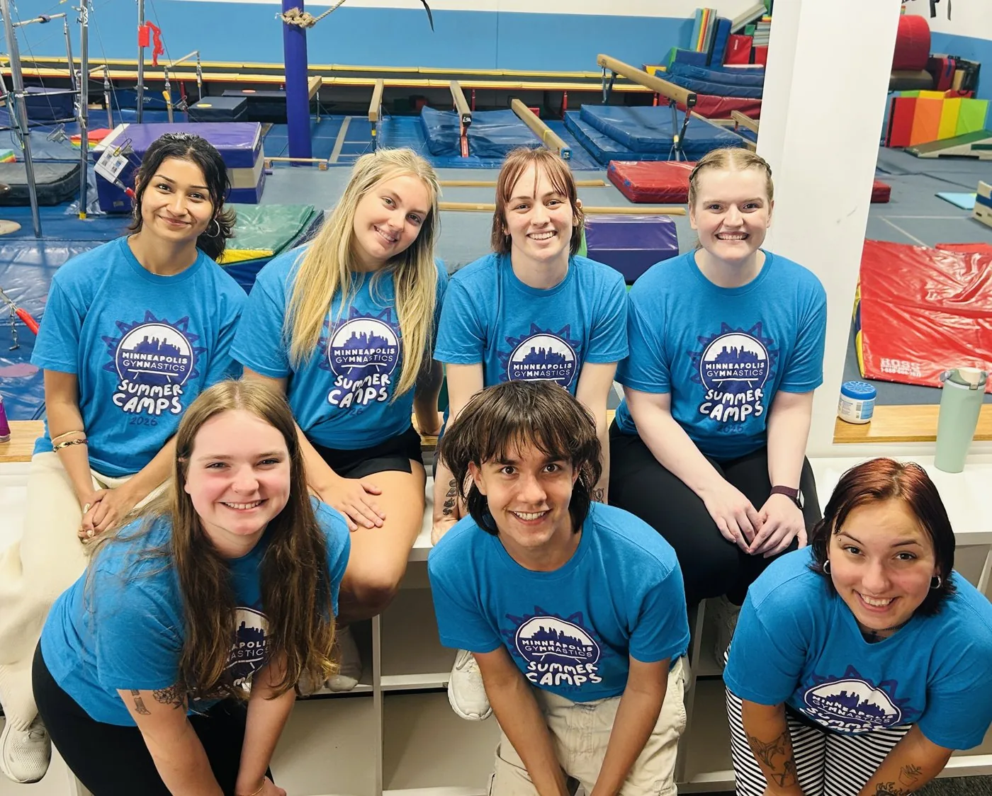 Seven summer camp coaches smile together in matching Minneapolis Gymnastics shirts