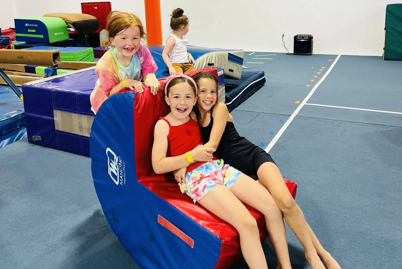 Three young gymnasts laugh together on a big red-and-blue rocker mat during party play time