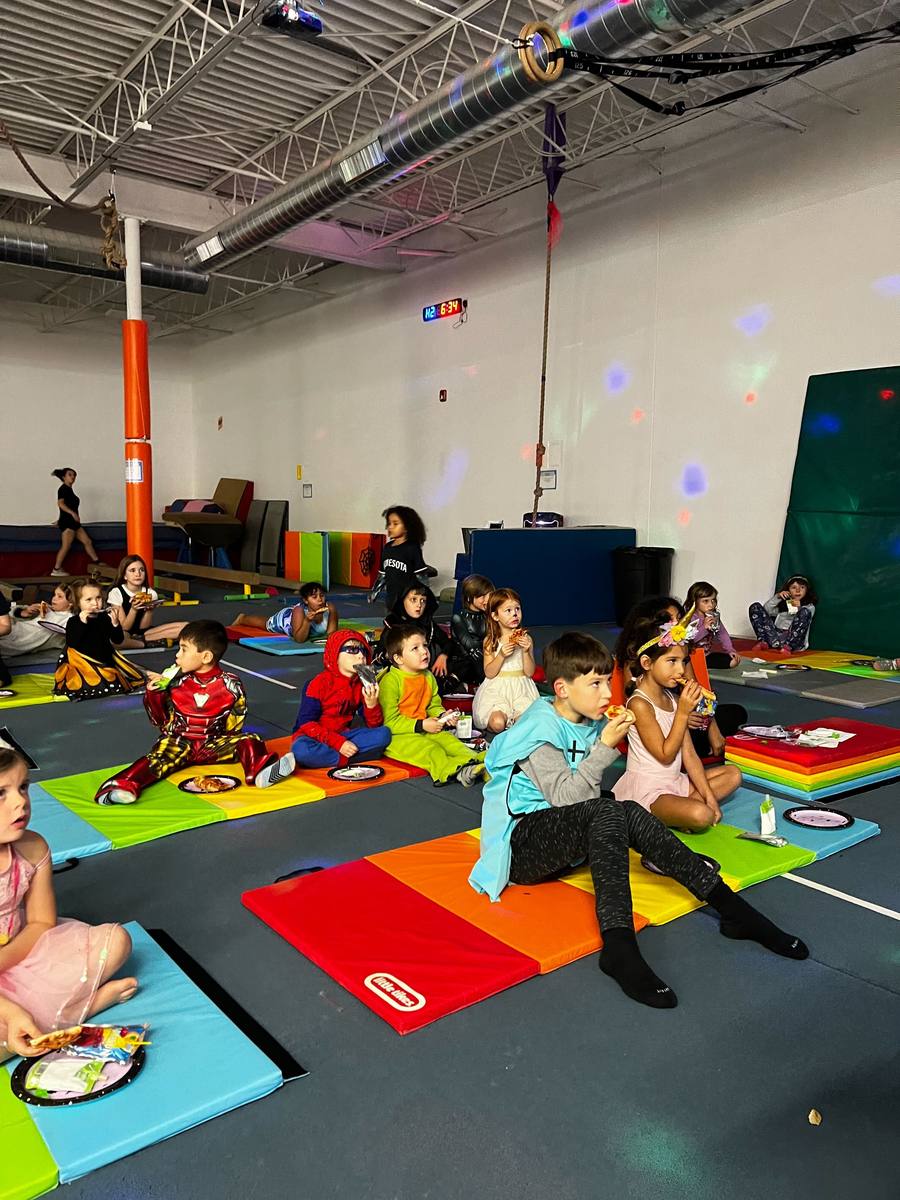 Kids in costumes eat pizza on colorful mats while watching a movie under party lights