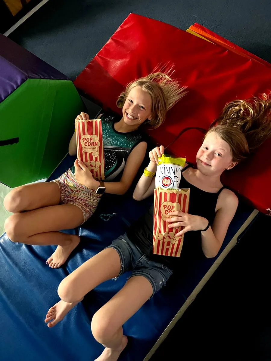Two girls grin with popcorn bags while lounging on mats at movie night